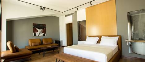 A modern hotel room with a large bed, a sofa, a bench, and a bathroom with a bathtub.