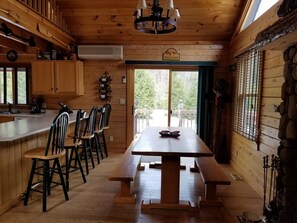 A wooden cabin interior with a dining table, chairs, a kitchen area, and a welcome sign. - Luxury Riverfront Log Cabin Retreat White Mountains, NH (Thornton)