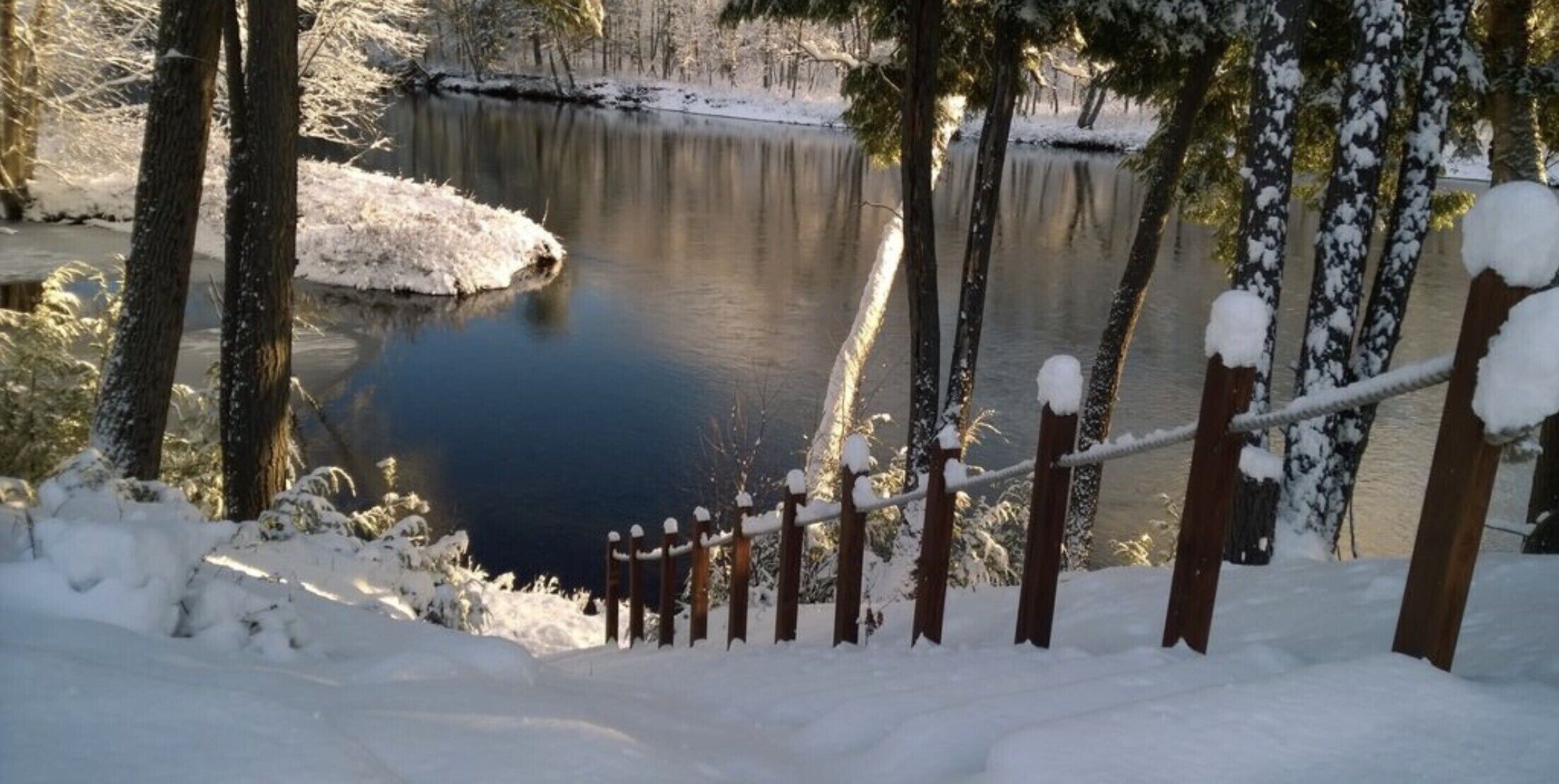A snow-covered wooden fence leading to a calm lake surrounded by trees.
