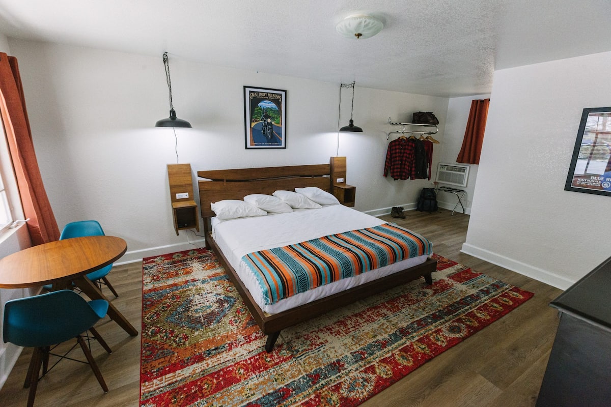 Ein Schlafzimmer mit einem hölzernen Bett, einem bunten Teppich, einem Schreibtisch mit Stuhl und einem kleinen Tisch.