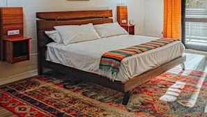 A bedroom with a wooden bed, a nightstand, a wall-mounted lamp, a framed picture, and a window with curtains. - Route 19 Inn (Maggie Valley)