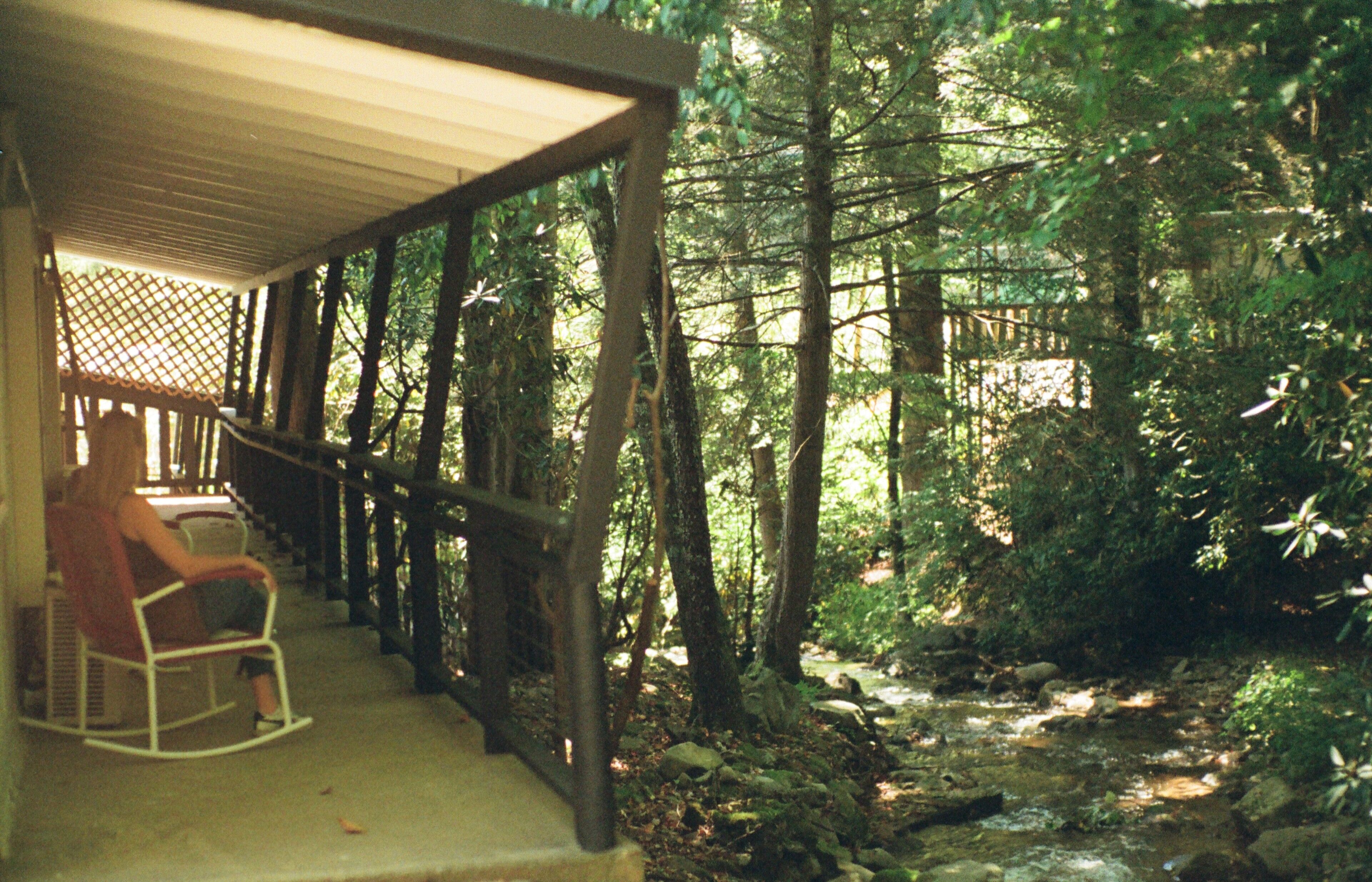 A wooden porch with a rocking chair, surrounded by a forested area and a stream.