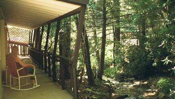 A wooden porch with a rocking chair, surrounded by a forested area and a stream.