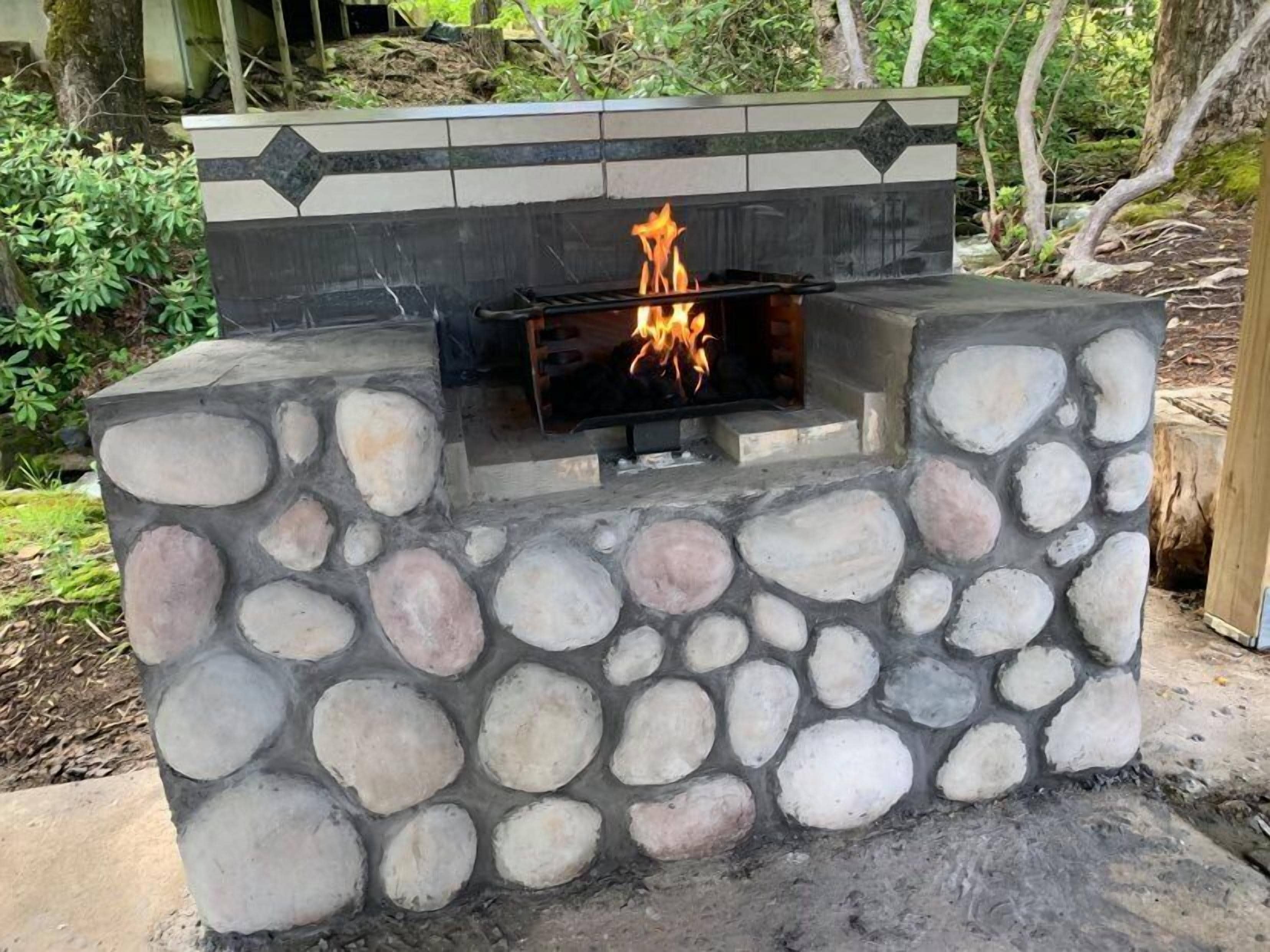 A stone-built outdoor fireplace with a fire burning inside, surrounded by greenery.