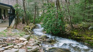 A rustic cabin with a porch and chairs overlooking a flowing stream in a forested area. - Route 19 Inn (Maggie Valley)
