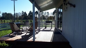 A covered outdoor deck with wooden chairs and a table, overlooking a fenced area with greenery.