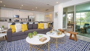 A modern living room with a grey sofa, yellow cushions, and a patterned floor. There is a kitchen in the background with white cabinets and a dining area with a glass door. - 8905 New Luxury Solara Resort Private Pool (Kissimmee)