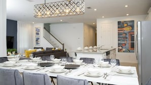 A modern dining area with a chandelier, white table set for six, and a kitchen in the background. - 8905 New Luxury Solara Resort Private Pool (Kissimmee)