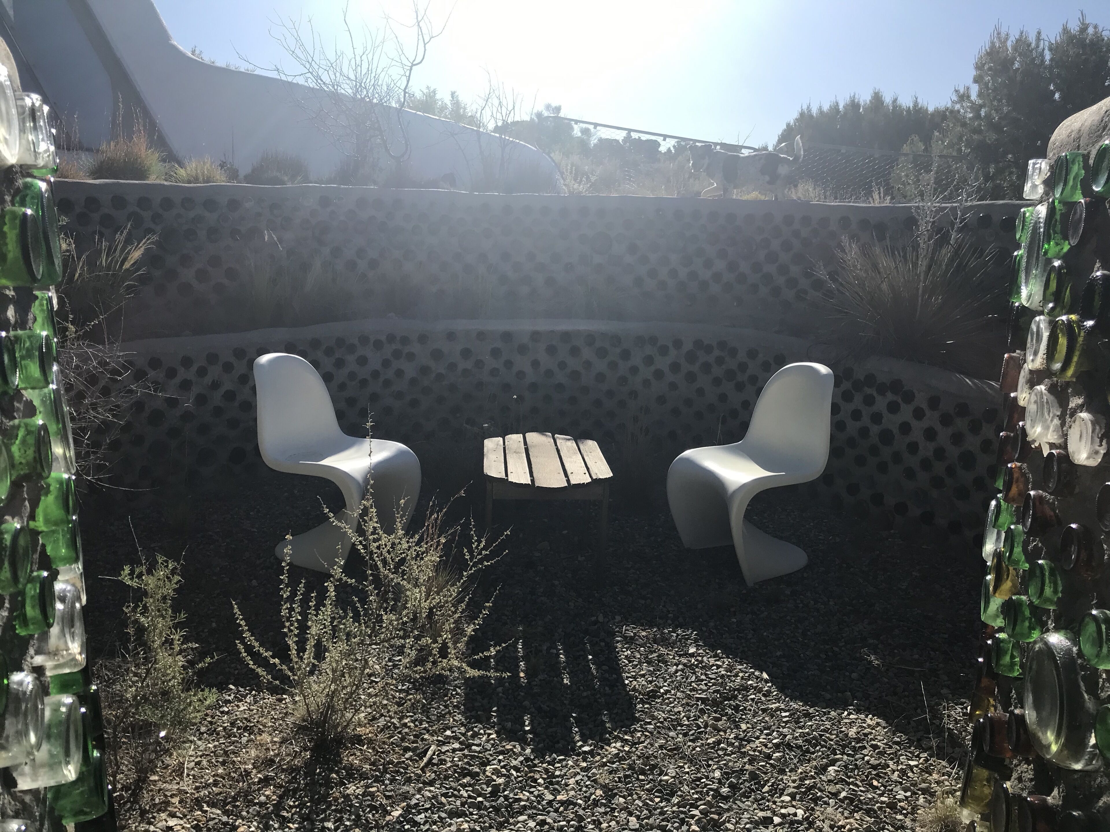 A patio area with two white chairs and a small table, surrounded by a wall decorated with green glass bottles.