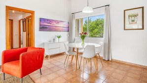 Un coin repas avec une table ronde, des chaises blanches et un fauteuil rouge. Il y a une fenêtre avec des rideaux, un tableau au mur et un petit meuble contenant des objets décoratifs.