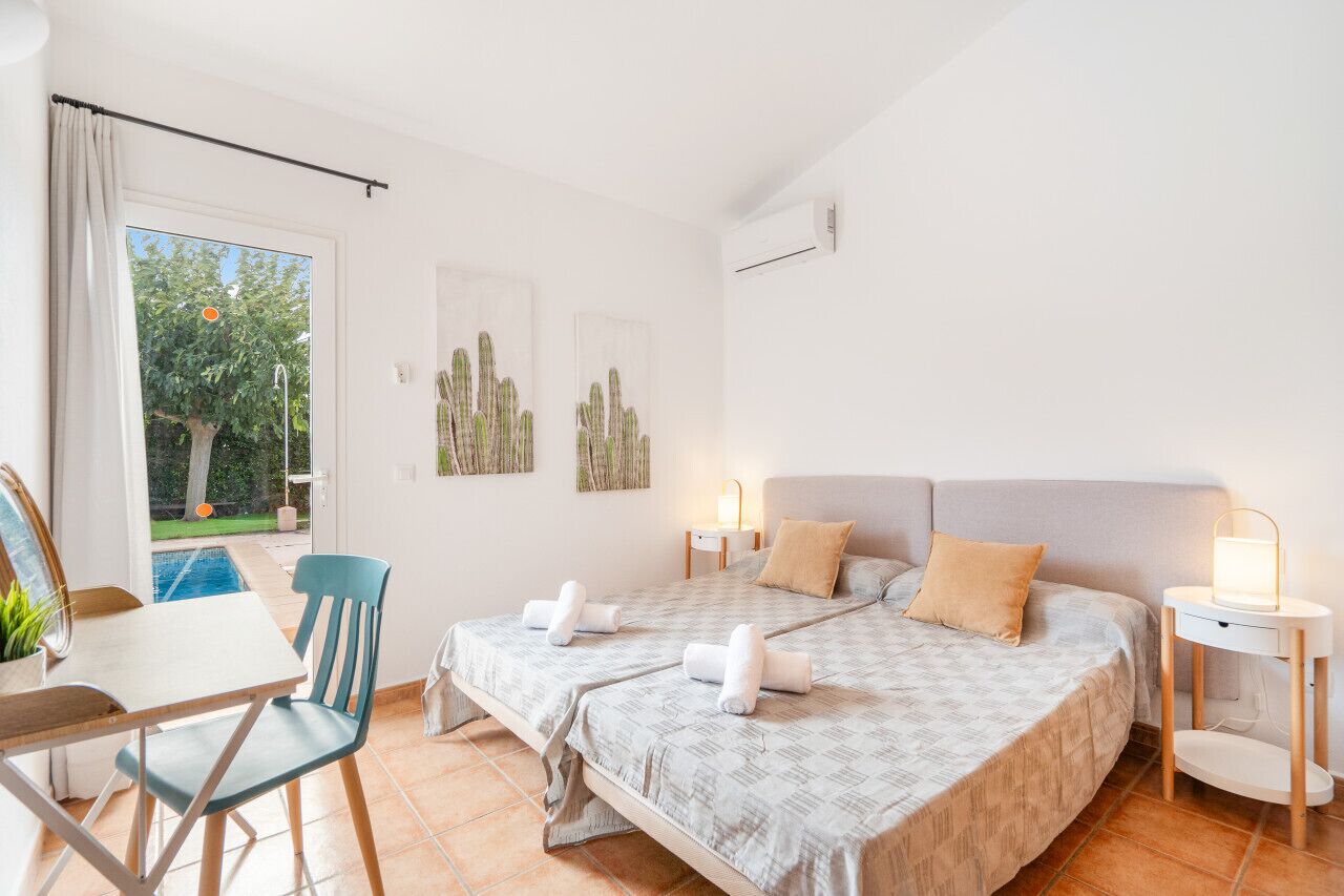 Un llit doble amb dos almoinos, una taula de nit amb una làmpada, una cadira, una taula petita amb una planta, i vista a un arbre i una piscina a l'exterior.