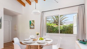 Un comedor con una mesa redonda dispuesta para cuatro, sillas blancas, una lámpara colgante blanca y una ventana que ofrece vistas a vegetación y un edificio.