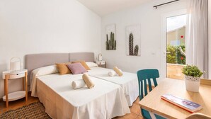 Habitación de hotel con dos camas, una mesita, una silla, un ventanal con cortinas y una planta en maceta.