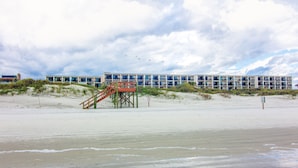 Ett hotell vid stranden med en trappa som leder upp till en terrass, omgivet av sanddyner och en molnig himmel.