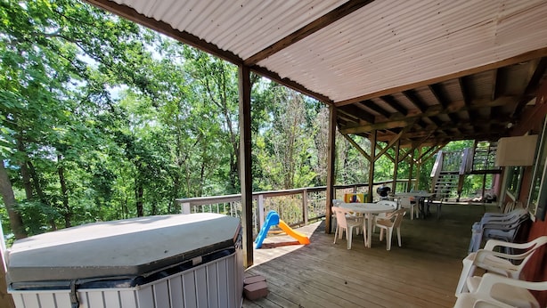 A covered outdoor deck with a hot tub, picnic tables, and chairs, surrounded by lush greenery. - Hog Heaven-Lower Level of Private Home- Lakefront (Garfield)