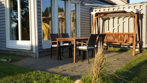 A patio with a wooden table and chairs, a swing set, and a sliding glass door leading to a room. - Beautiful vacation home in a quiet location (Mönchneversdorf)