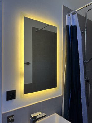 A modern bathroom with a large mirror, a shower curtain, and a visible shower head. - Small studio apartment by the park with parking space (Lüneburg)