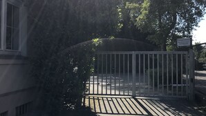 Een hekpoort met een metalen omheining, een verharde oprit en een gebouw met ramen aan de linkerkant.