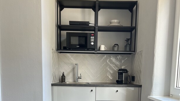 Een functionele keuken met een magnetron, koffiezetapparaat en een klein kastje. Een ronde tafel en een stoel met een kussen in vorm van een hert staan op de voorgrond.