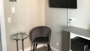 Un saló modern amb una butaca gris, una taula de vidre petita, un televisor de pantalla plana muntat a la paret i un moble blanc per allotjar aparells electrònics.