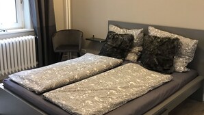 Un dormitori modern amb llit gris, almoinades estampades, una taula de nit i una làmpada penjada a la paret.