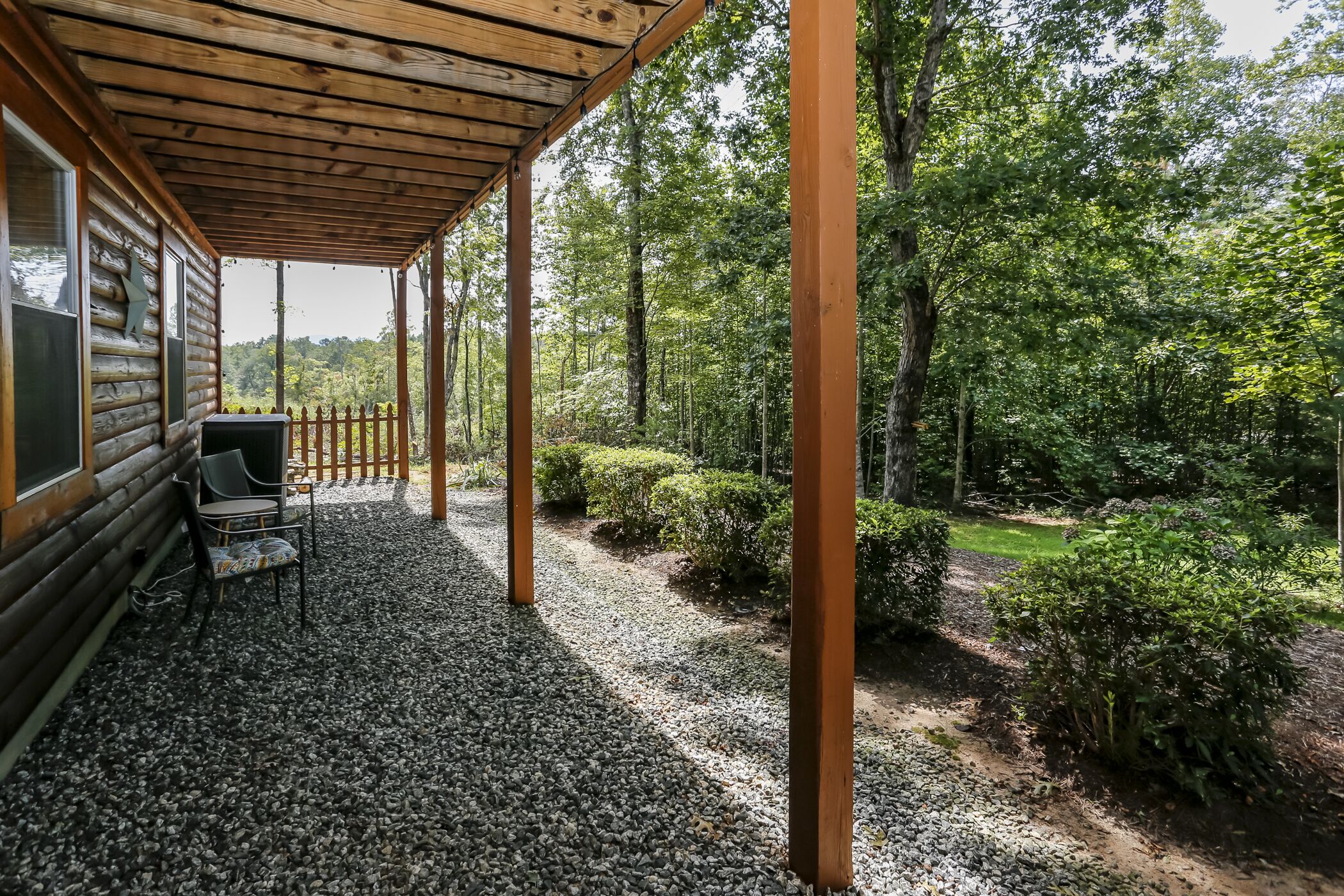 Une cabane en bois dotée d’un porche couvert, d’un sol en gravier et offrant une vue sur une zone boisée.