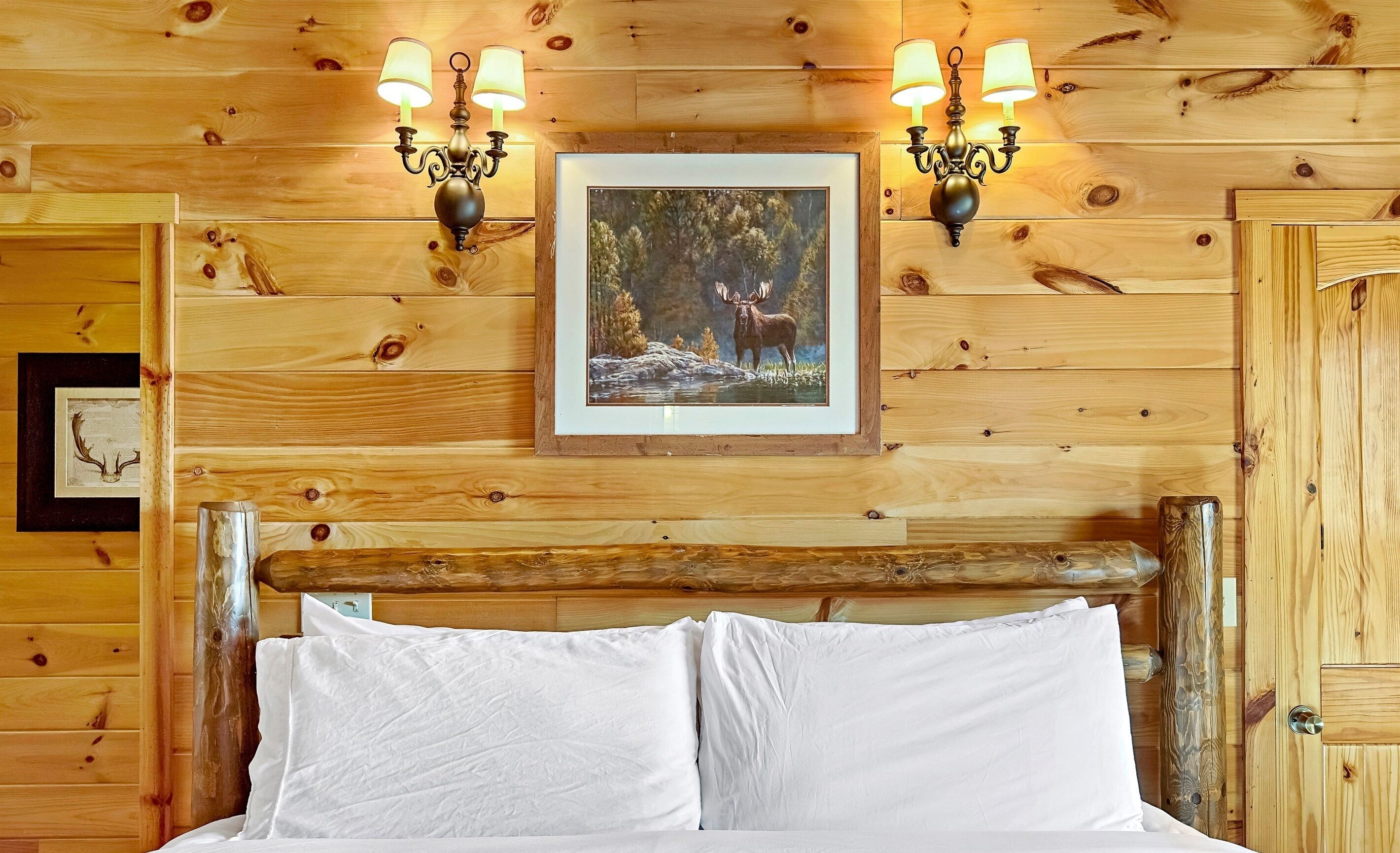Una habitación de cabaña de madera con una cama, dos lámparas de pared y un cuadro enmarcado de un alce.