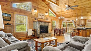 A cozy cabin interior with a stone fireplace, wooden ceiling, and large windows offering views of the outdoors.