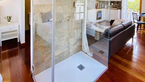 A modern bathroom with a glass shower enclosure, a wooden ceiling, and a visible kitchen area with white cabinets.