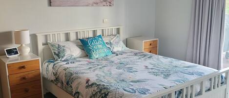 A bedroom with a bed, bedside tables, a ceiling fan, a beach-themed painting, and a window with curtains.