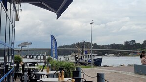 An outdoor seating area with tables and chairs, a blue awning, and a view of the river with boats and a bridge in the background. - Catalina Coastal Getaway / Family friendly home (Catalina)