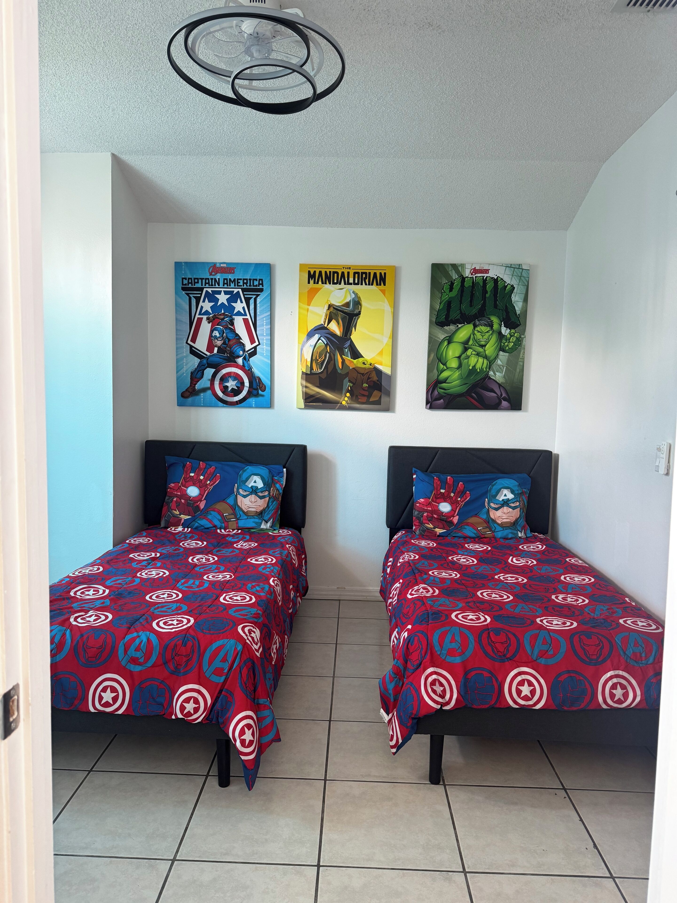 A bedroom with two beds, each with a Captain America themed bedspread. There are three posters on the wall: Captain America, The Mandalorian, and Hulk.