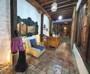 A narrow corridor with a sofa and a mannequin in the foreground, wooden beams on the ceiling, and a brick wall on one side. - Hotel El Zaguán Histórico (San Cristóbal de las Casas)