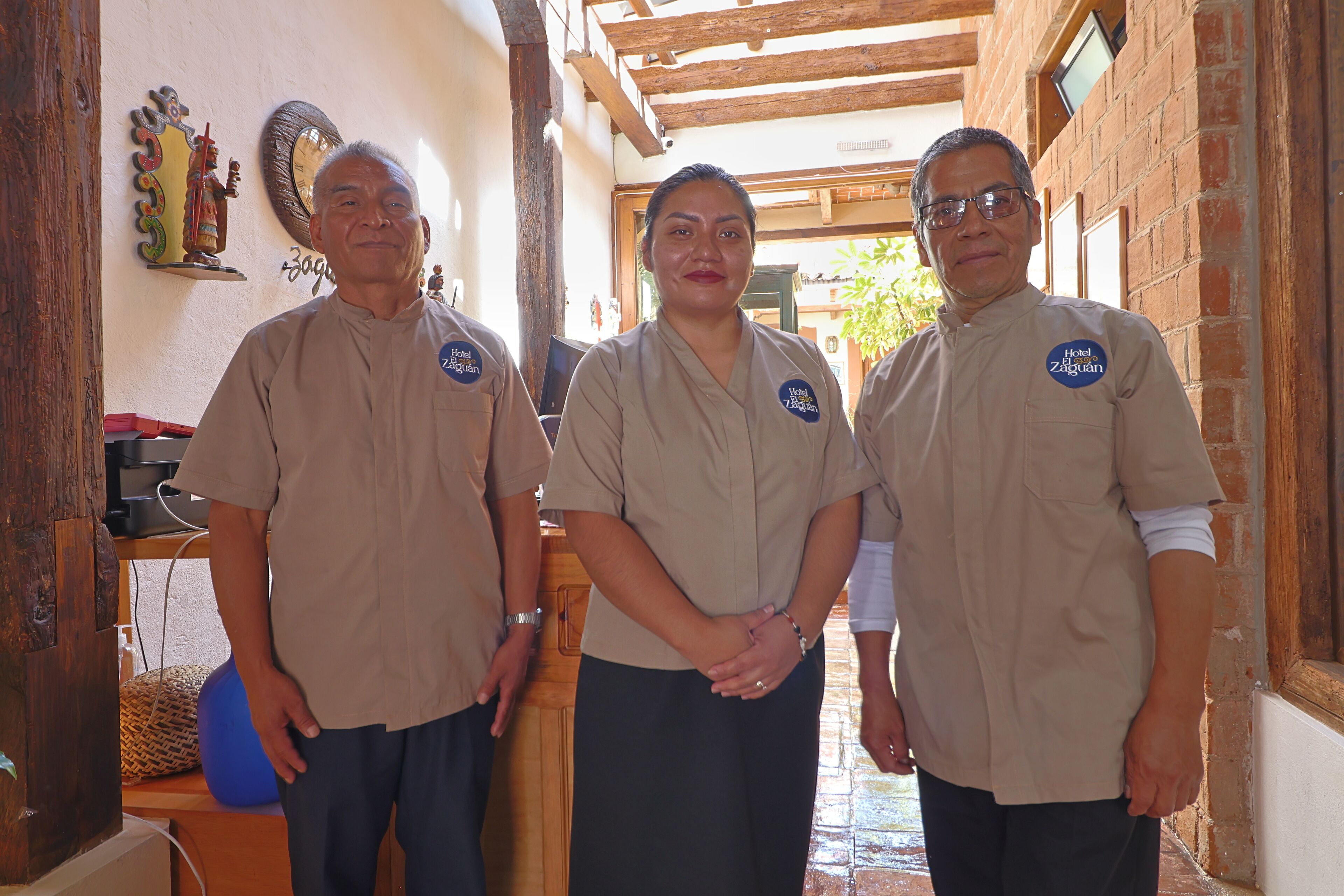 Tres personas en un pasillo con techo de madera y paredes de ladrillo, vestidas con uniformes que incluyen una insignia que dice 'Hotel Záhuán'.