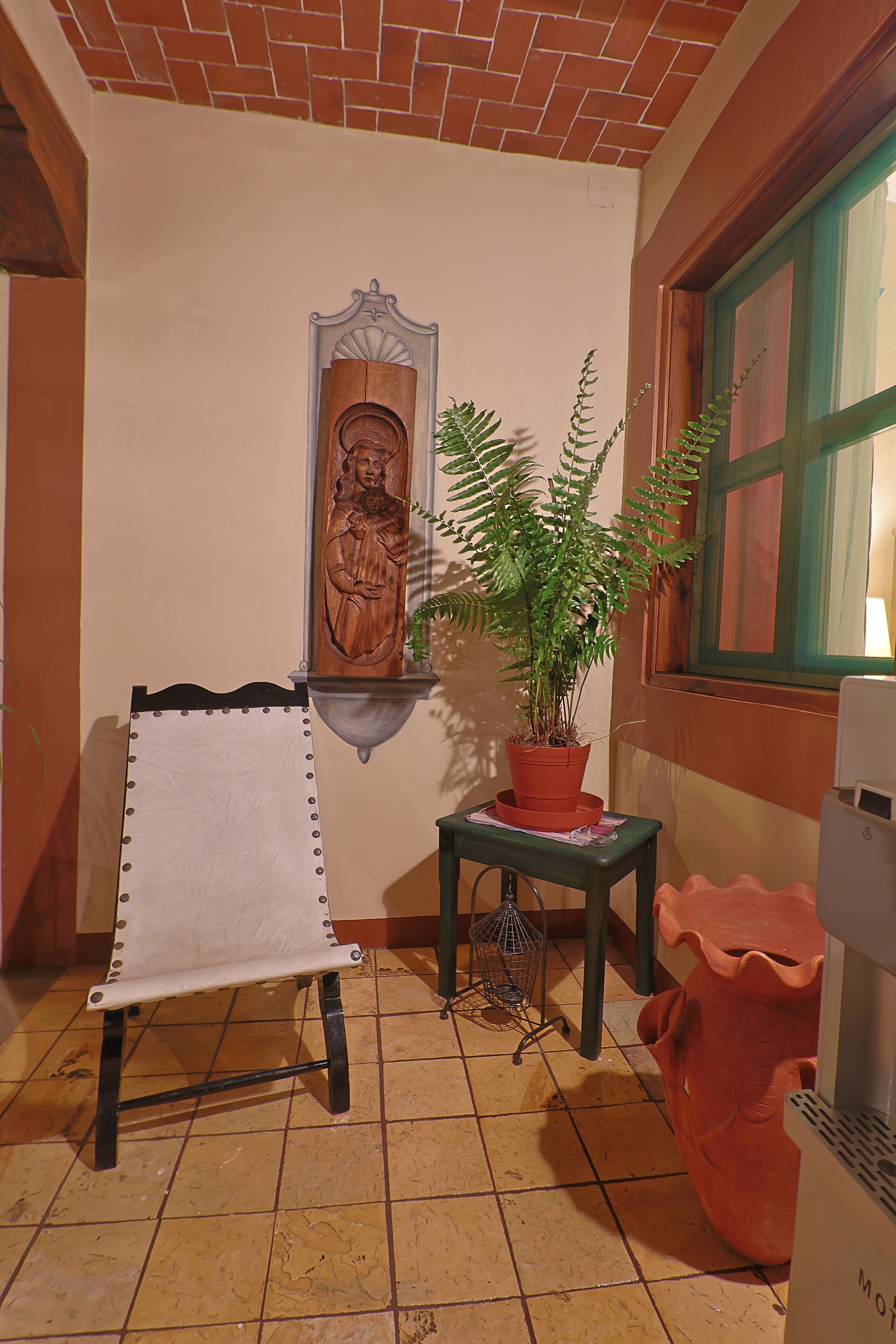 A room with a wooden bench, a potted plant, a wall-mounted sculpture, and a window with a green frame.