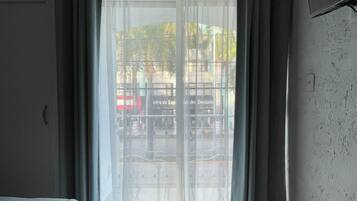 Habitación de hotel con cama, mesita de noche de madera, ventana con cortinas translúcidas y un equipo de aire acondicionado en la pared.