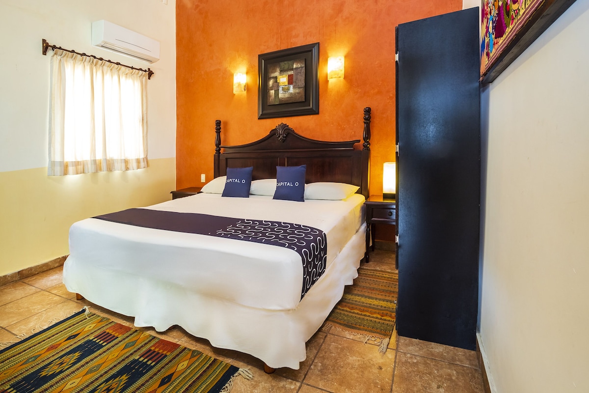A hotel room with a large bed, a headboard, a wall-mounted air conditioner, a window with curtains, and a patterned rug.