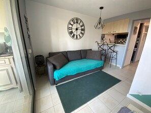 Een woonkamer met een bank, een groen vloerkleed, een wandklok en een keuken op de achtergrond.