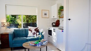 A modern living room with a blue sofa, a coffee table with books and a vase, a kitchen with white cabinets, and a window with a view of greenery. - Beach, Cafes, Golf, Sun & Privacy at Paraparaumu Beachside Studio w sea view (Paraparaumu)
