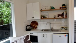 A compact kitchen with white cabinets, a sink, and a refrigerator. There are shelves above the counter holding various items. - Beach, Cafes, Golf, Sun & Privacy at Paraparaumu Beachside Studio w sea view (Paraparaumu)