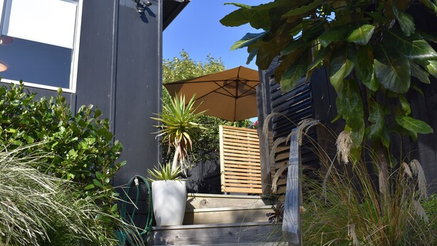 A wooden staircase leading to a house with a dark exterior, a large umbrella, and various plants. - Beach, Cafes, Golf, Sun & Privacy at Paraparaumu Beachside Studio w sea view (Paraparaumu)