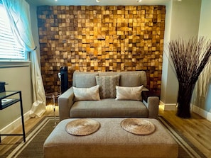 Una sala de estar con un sofá, dos cojines, una mesa de centro con un camino de mesa tejido, una pared con diseño de bloques de madera y un jarrón alto con ramas.