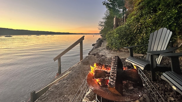 A waterfront deck with a fire pit, Adirondack chairs, and a railing overlooking calm water.