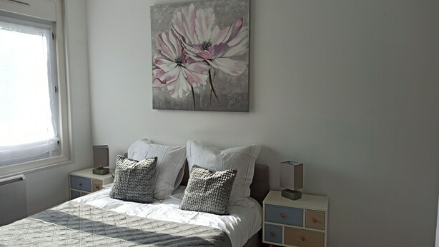 A bedroom with a bed, pillows, a bedside table, a lamp, a nightstand, a painting, and a window. - Les Chevalets II/ University/Louvre/Bollaert (Lens)