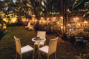 An outdoor seating area with wicker chairs and a round table, surrounded by lush greenery and string lights. - Winner Inn (Yangon)