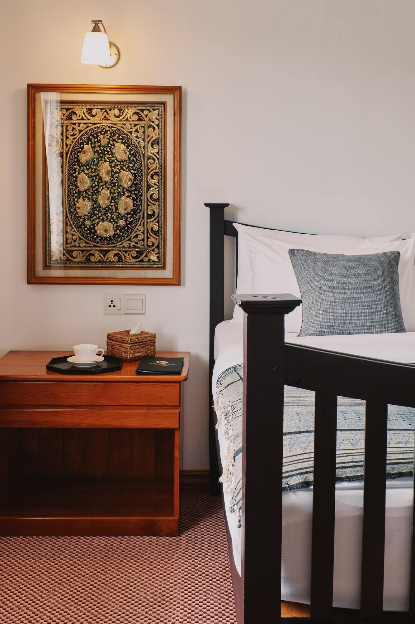 A bedroom with a bed, a bedside table with a cup and a book, a wall-mounted lamp, and a framed artwork above the bed.