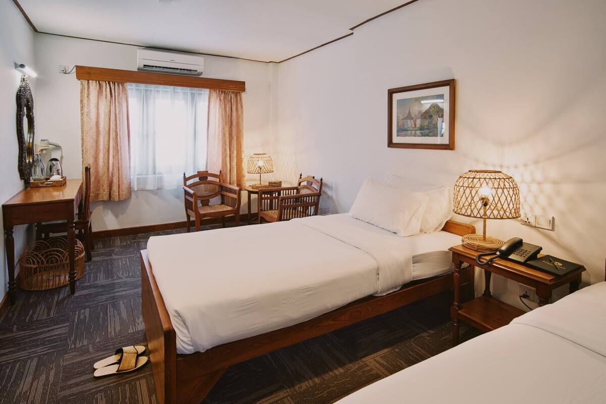 A hotel room with two beds, a desk with a chair, a table with chairs, a lamp, and a framed picture on the wall.