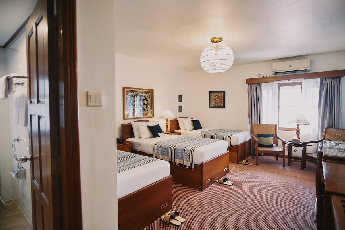 A hotel room with two beds, a sitting area with a chair and table, a hanging light fixture, and a window with curtains.