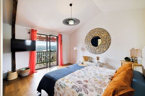 A bedroom with a bed, a television, a balcony with a view, and a round mirror on the wall. - Spacious BAÏGURA studio, mountain views, direct access to pool & spa (Hasparren)