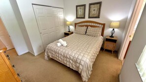 A bedroom with a bed, two bedside tables, a nightstand, a door, and two wall-mounted pictures.